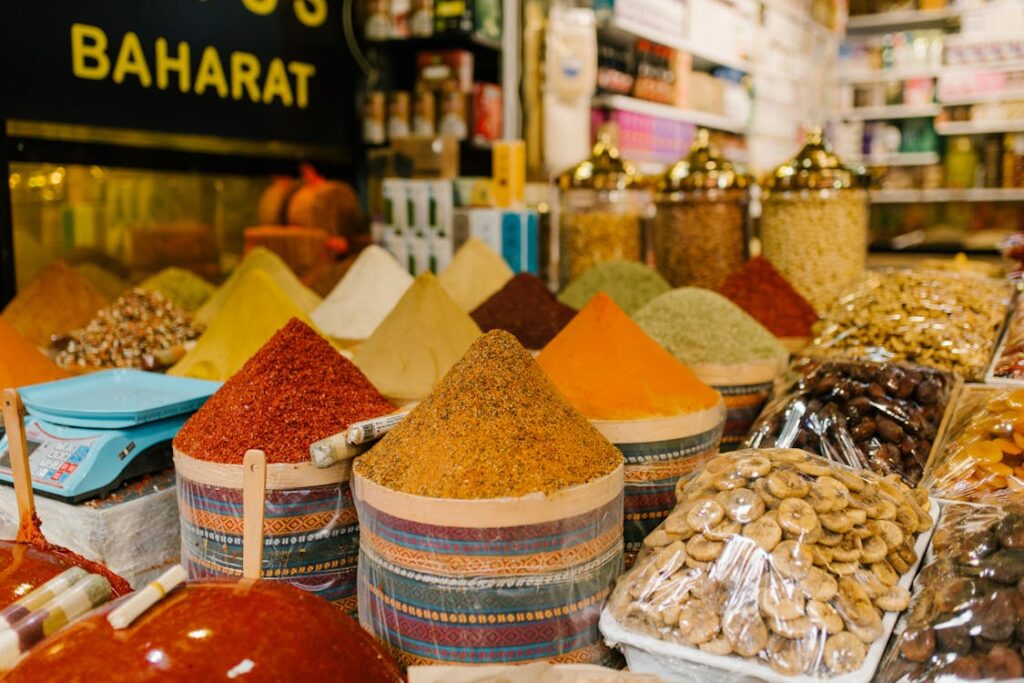 Photo d'étalage d'épices colorées dans un souk marocain, avec des tas d'épices en poudre de différentes couleurs (rouge, jaune, orange, vert) disposés dans des paniers, et des fruits secs emballés en arrière-plan. Panneau 'BAHARAT' visible en haut à gauche.