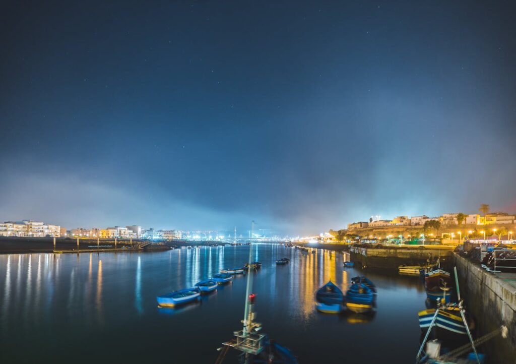 Une scène nocturne paisible d'un port, avec des bateaux sombres flottant sur une eau calme et les lumières scintillantes de la ville en arrière-plan