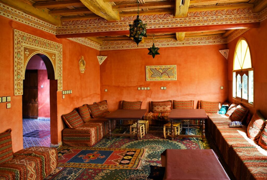 Intérieur d'un salon marocain traditionnel avec des murs orange texturés, des banquettes basses avec des coussins à motifs, et des tables basses. Le plafond en bois apparent est décoré de motifs géométriques et une lanterne en métal est suspendue. Une porte cintrée mène à une autre pièce. Des tapis colorés recouvrent le sol.