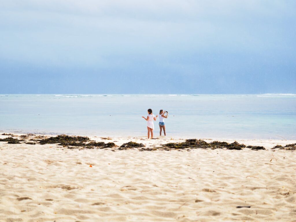 Photo de deux personnes sur une plage de sable blanc avec des algues éparses, face à une mer calme aux eaux turquoise, sous un ciel bleu clair et légèrement nuageux.