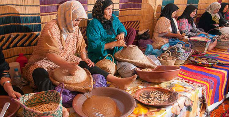 Photo de femmes assises dans une tente traditionnelle marocaine, travaillant à la préparation de l'huile d'argan avec des paniers, des plats en terre cuite et des noix, sur une table colorée avec un fond en nattes tressées.