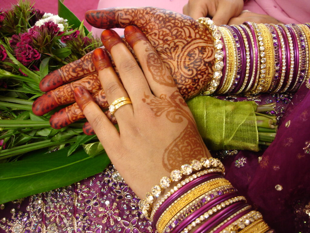 Photo de mains ornées de motifs au henné, portant des bracelets colorés et des bagues en or, posées sur un tissu violet scintillant avec un bouquet de fleurs roses à côté.