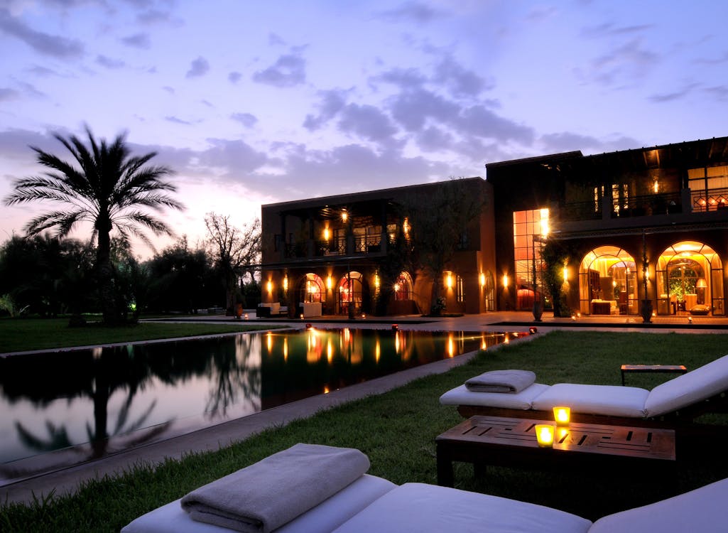Image d'un riad de luxe au crépuscule. On voit une piscine sombre reflétant les lumières du bâtiment et le ciel nuageux. Deux chaises longues avec des serviettes pliées sont au premier plan, et un palmier se dresse à gauche. Le bâtiment en arrière-plan a des arches éclairées et des fenêtres, suggérant une atmosphère chaleureuse