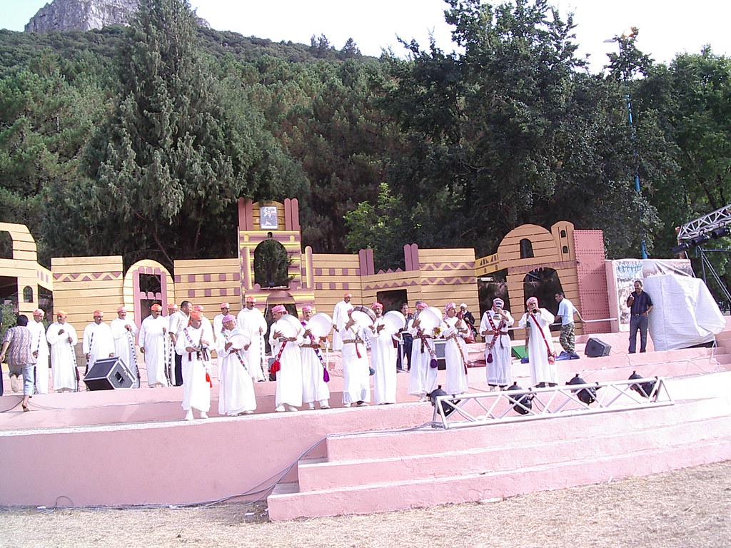 Photo d'une représentation traditionnelle marocaine en plein air, avec des musiciens en tenues blanches sur une scène décorée, jouant des instruments comme des tambours, entourés d'arbres et de collines.