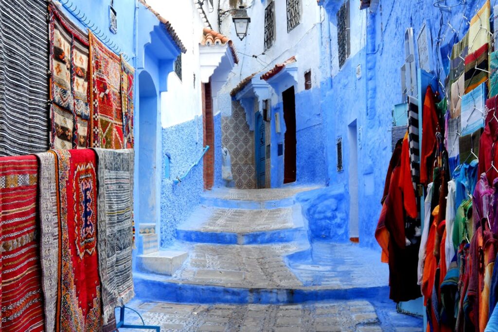 Ruelle pavée de Chefchaouen bordée de maisons aux murs bleus et blancs, avec des escaliers menant à une porte ornée de motifs. Des tapis et des vêtements colorés sont suspendus de chaque côté de la ruelle.