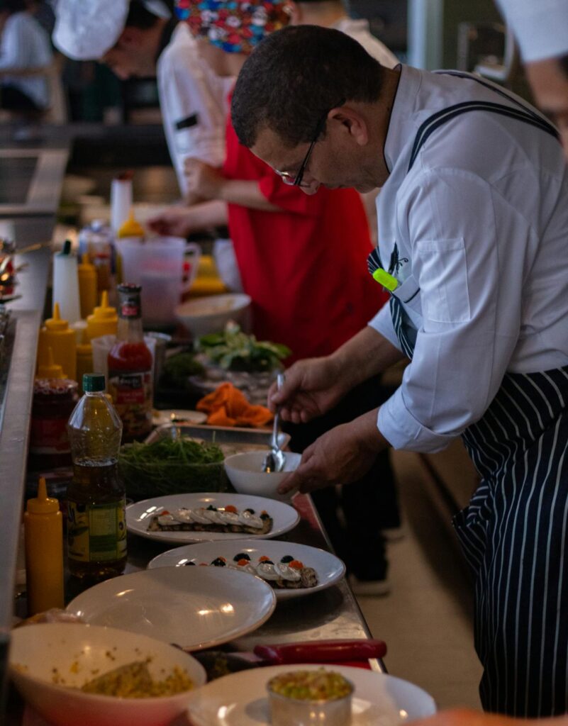 Photo d'un chef cuisinier en tablier rayé préparant des plats sur un comptoir de cuisine, avec des assiettes de nourriture, des bouteilles d'assaisonnements et des légumes frais autour, dans une ambiance de travail en cuisine professionnelle.