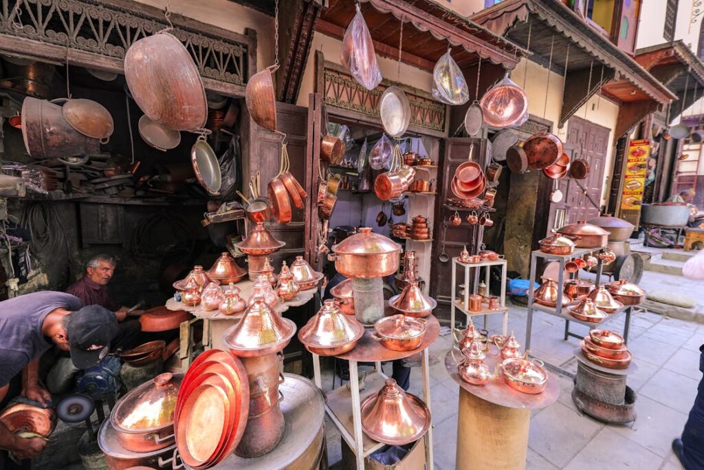 Une boutique d'artisanat traditionnelle avec des ustensiles en cuivre suspendus et exposés. On voit des tagines en cuivre brillant sur des étagères, ainsi que des casseroles et des plats suspendus. Deux artisans travaillent à l'arrière-plan, l'un assis et l'autre debout, dans ce qui semble être un atelier ou une rue commerçante.