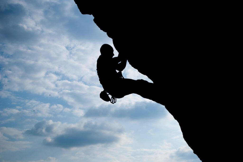 Photo en silhouette d'une personne escaladant une falaise sous un ciel bleu avec des nuages, équipée de matériel d'escalade, dans un paysage naturel.