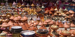 Un étalage coloré de poteries artisanales dans un marché. On voit une multitude de tagines en terre cuite de différentes tailles, ainsi que des assiettes et d'autres objets décoratifs. Les couleurs dominantes sont le marron de la terre cuite et les motifs bleus et blancs des assiettes.