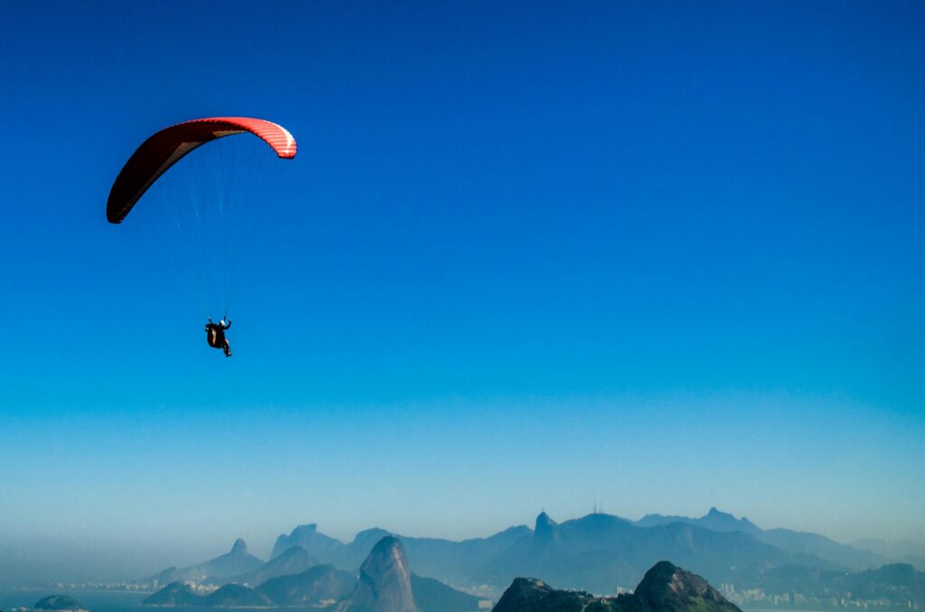 Photo d'une personne en parapente avec une voile rouge, survolant un paysage montagneux sous un ciel bleu clair, avec des sommets et une ville visibles en arrière-plan