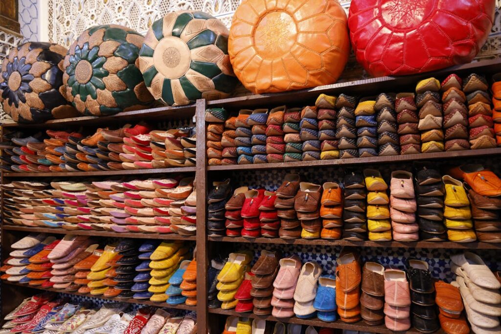 Étalage coloré de babouches et poufs marocains dans une boutique. Les babouches, de différentes couleurs et styles, sont rangées sur plusieurs étagères en bois. Au-dessus, des poufs en cuir de formes et couleurs variées, dont un rouge, un orange et un vert à motifs, sont disposés.