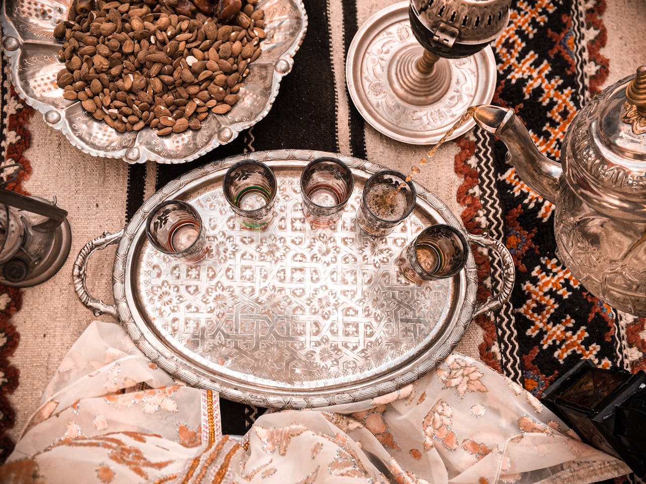 Une scène traditionnelle avec un plateau en argent orné de motifs complexes, contenant six petits verres, dont certains remplis de thé. Autour du plateau, sur une couverture tissée aux motifs géométriques rouges et noirs, se trouvent une théière en métal gravée versant du thé, un bol en argent rempli de noix, un narguilé en métal, et un tissu léger aux tons beige et rose pâle. L'ambiance évoque une hospitalité chaleureuse et culturelle.