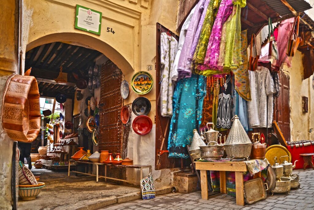 Photo d'une boutique dans un souk marocain, avec des étals remplis de poteries, de plats colorés, de vêtements traditionnels suspendus et de tajines en métal, dans une ruelle pavée d'une médina