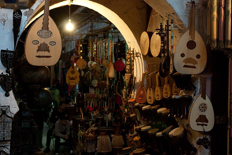 Photo d'une boutique d'instruments de musique traditionnels dans un souk, avec des luths suspendus, des tambours empilés et d'autres instruments à cordes visibles. Un homme est assis à l'arrière-plan. L'éclairage est doux et chaud.