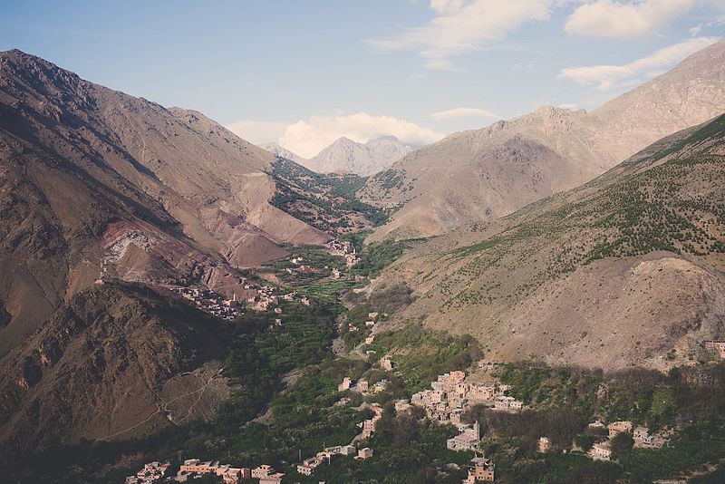 Photo d'une vallée encaissée dans les montagnes de l'Atlas, avec un petit village niché au centre, entouré de collines rocailleuses et de verdure, sous un ciel bleu avec quelques nuages.