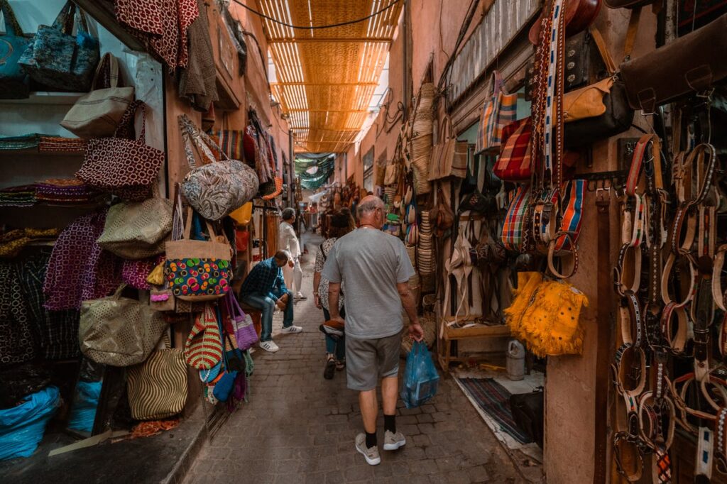 Photo d'une ruelle animée d'un souk marocain, avec des visiteurs parcourant des étals remplis de sacs colorés, de textiles et de ceintures artisanales, sous un toit en lattes de bois