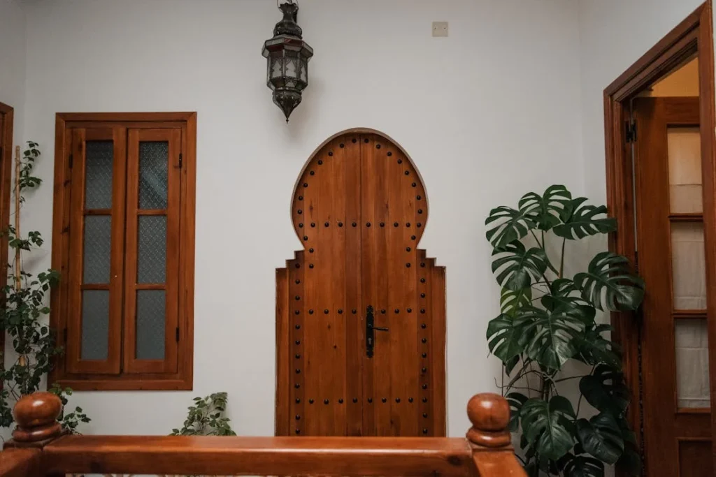 Vue intérieure d'un bâtiment avec une porte en bois sculpté de style marocain au centre, flanquée d'une fenêtre en bois à gauche et d'une porte ouverte à droite. Une plante verte est visible à droite, et une lanterne suspendue au plafond. La perspective est légèrement en contre-plongée, avec une rampe d'escalier en bois visible en bas de l'image.