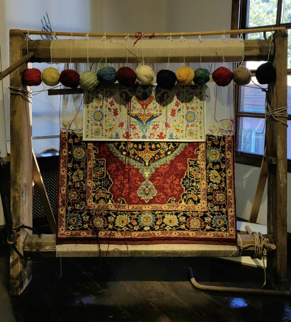 Métier à tisser traditionnel en bois avec un tapis en cours de fabrication. Le tapis présente des motifs floraux complexes dans des tons de rouge, bleu et jaune. Des pelotes de laine colorées sont suspendues au-dessus du métier, prêtes à être utilisées. Le métier est installé dans une pièce avec une fenêtre à l'arrière-plan.