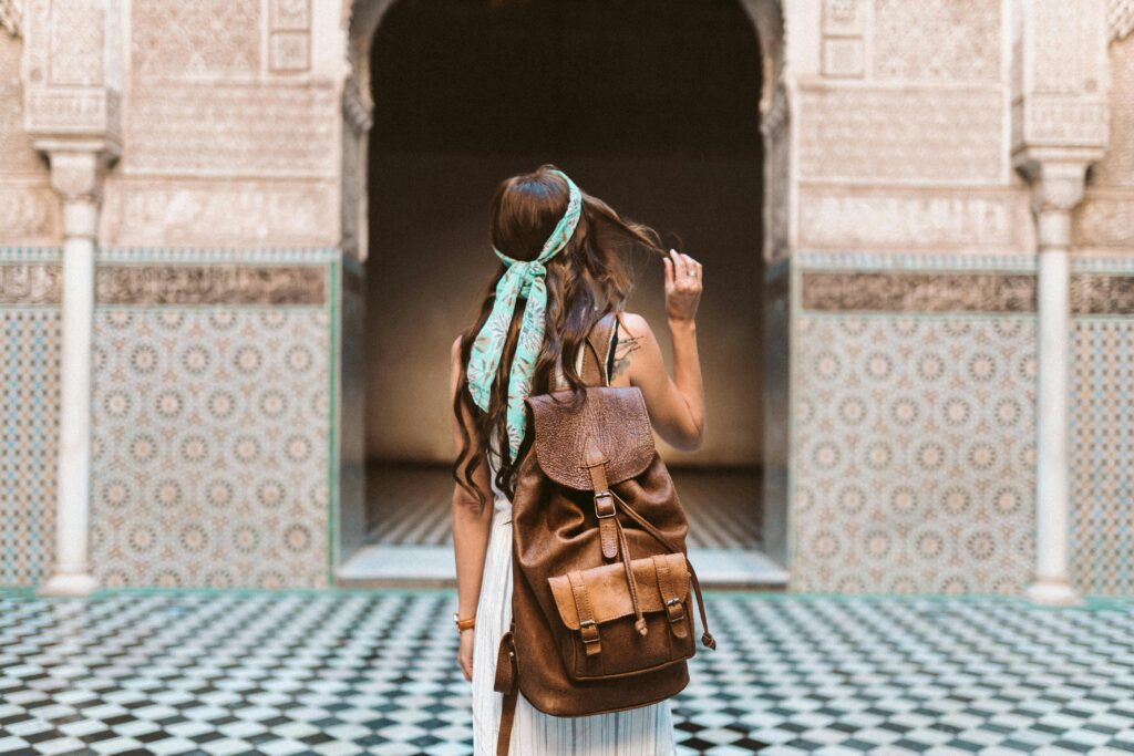 Photo d'une femme de dos portant un sac à dos en cuir marron et un foulard vert, se tenant dans un patio marocain aux murs ornés de carreaux géométriques et à l'architecture traditionnelle avec arches.