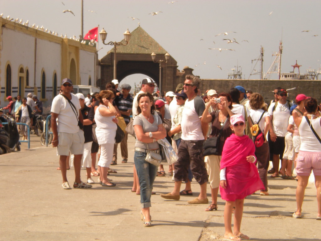 Photo d'un groupe de touristes rassemblés près d'une entrée architecturale marocaine avec un arc, des mouettes volent au-dessus, des bateaux visibles en arrière-plan et un bâtiment coloré à gauche