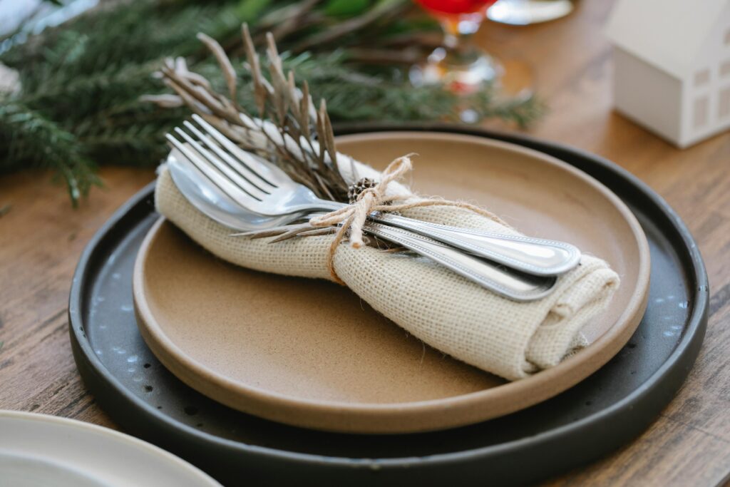 "Une élégante mise en place de table avec une assiette beige sur une assiette noire, décorée d'un rouleau de serviette en tissu beige entourant des couverts (deux fourchettes et une cuillère) attachés avec une cordelette rustique. Des brindilles sèches et des branches de sapin vert ajoutent une touche naturelle à l'ensemble, posé sur une table en bois. Un petit objet décoratif en forme de maison blanche et un verre coloré sont visibles en arrière-plan."