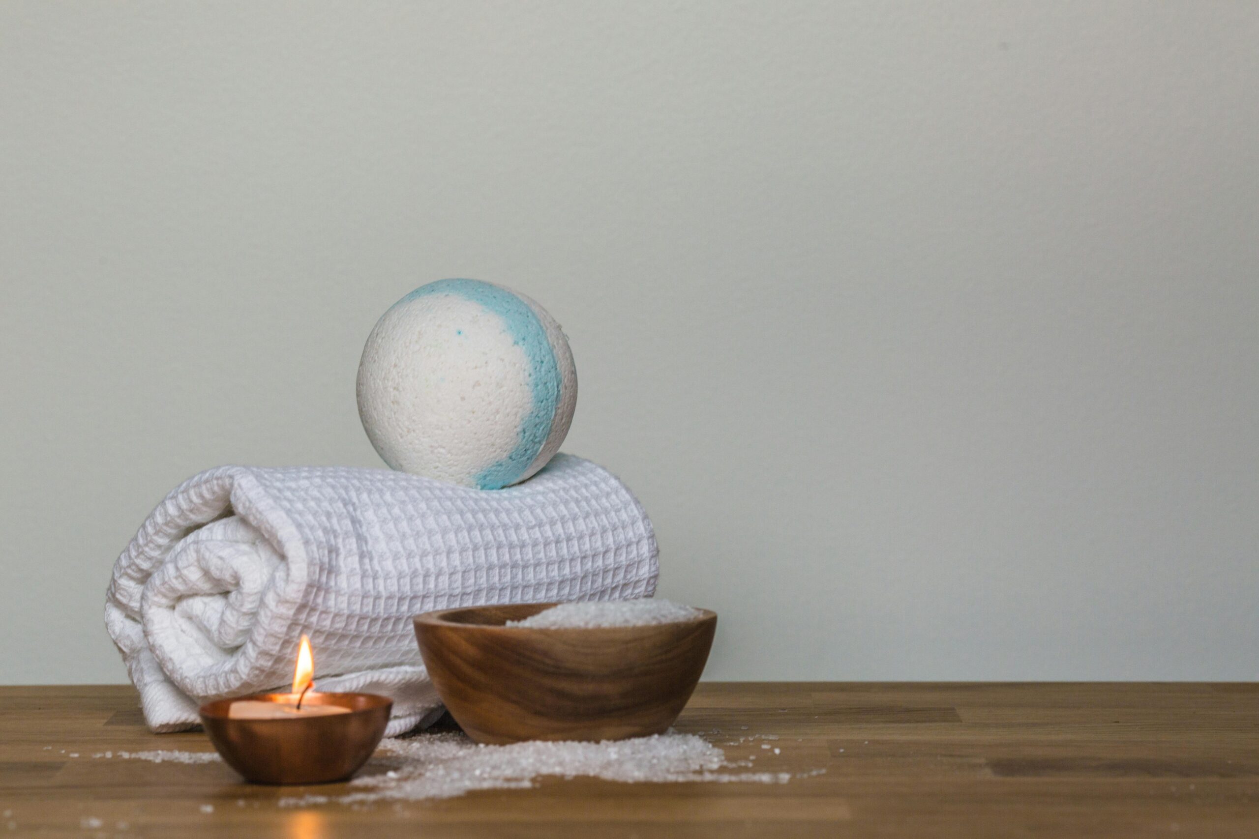 Photo d'une scène de spa avec une serviette blanche roulée, une bombe de bain blanche et bleue, un bol en bois rempli de sels de bain et une bougie allumée sur une table en bois clair.