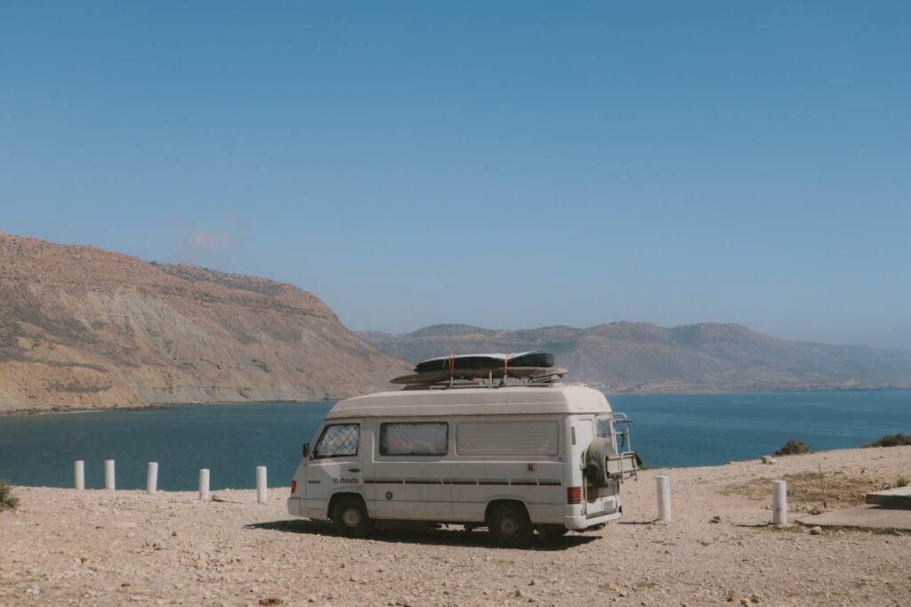 Un camping-car vintage blanc est stationné sur une falaise surplombant une mer d'un bleu profond, avec des montagnes arides en arrière-plan. Des planches sont attachées sur le toit du véhicule, évoquant un mode de vie nomade et l'appel de l'aventure