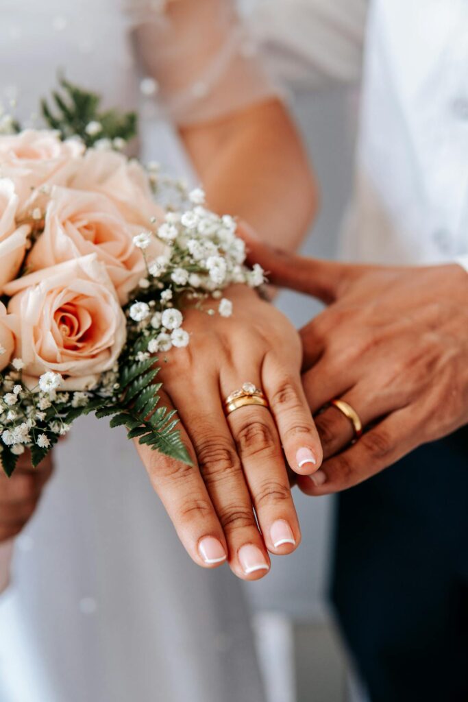 Close-up de deux mains tenant un bouquet de mariage avec des roses roses et de la verdure, orné de petites fleurs blanches. Les mains portent des alliances en or, symbolisant l'union lors d'une cérémonie de mariage, avec un fond flou montrant des vêtements blancs et sombres.