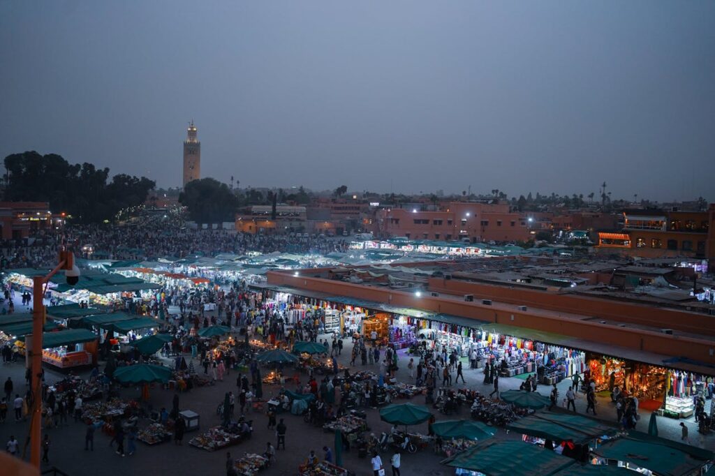 Vue panoramique d'un marché animé au crépuscule à Marrakech, avec de nombreux étals sous des toits verts, une foule de personnes, et la silhouette illuminée de la Koutoubia en arrière-plan sous un ciel sombre.