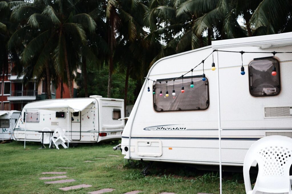 Photo d'un camping avec deux caravanes blanches, décorées de guirlandes lumineuses colorées, stationnées sur une pelouse verte. Des chaises blanches et une table sont visibles à côté, avec des palmiers en arrière-plan et un bâtiment coloré au loin.