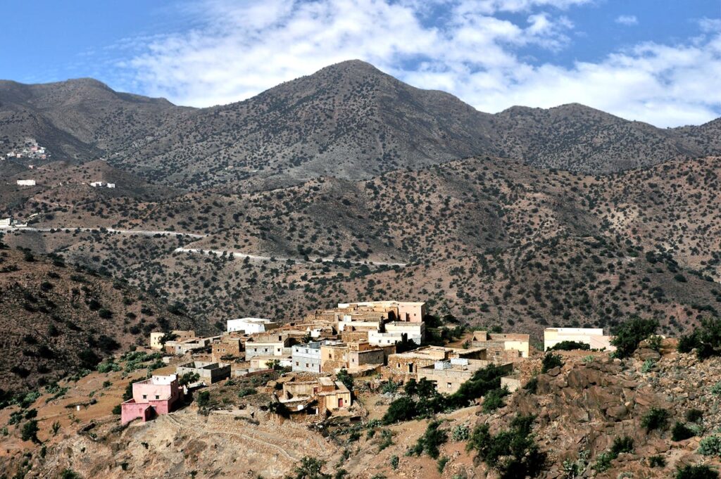 Vue panoramique d'un village marocain niché dans une vallée montagneuse aride, entouré de collines brunes et de terrasses agricoles. Un arbre vert et des rochers dominent le premier plan, avec une route sinueuse en arrière-plan