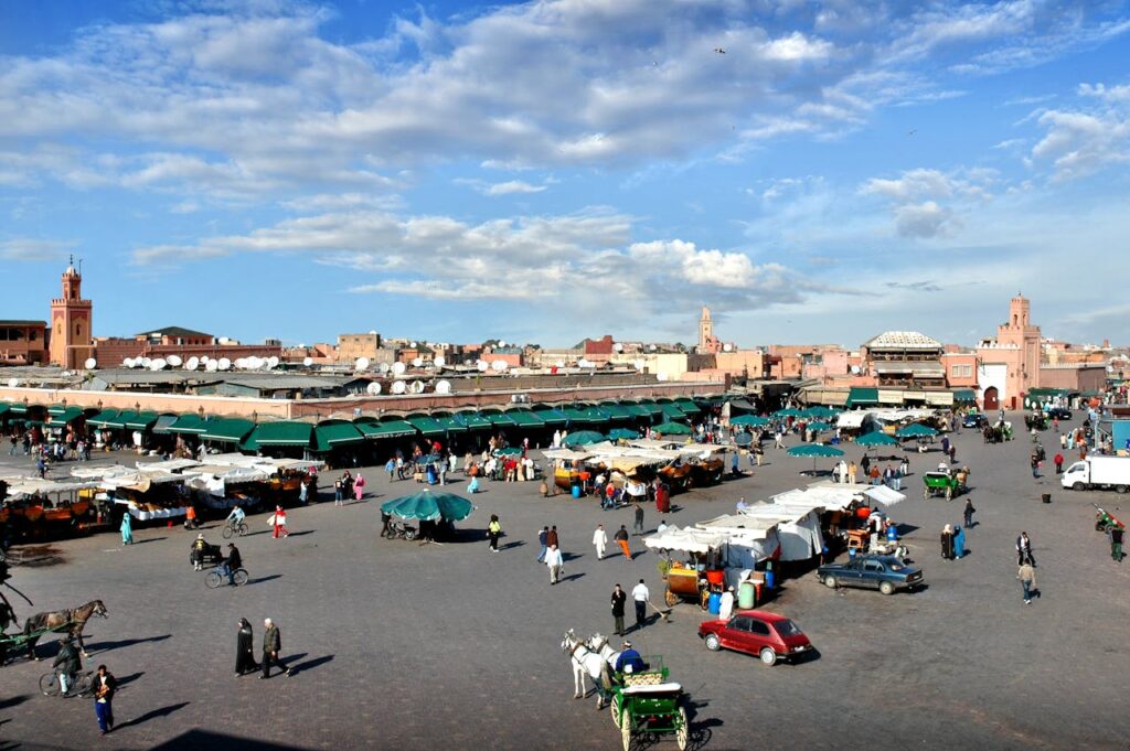 Vue panoramique de la place Jemaa el-Fna à Marrakech sous un ciel bleu nuageux, avec des étals de marché couverts, des charrettes à cheval, des passants, et des bâtiments ocre en arrière-plan, dont un minaret.