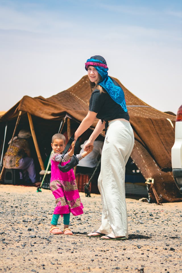 Une femme souriante portant un turban bleu joue avec une petite fille vêtue d'une robe traditionnelle rose dans un décor désertique. Derrière elles, une tente berbère abrite des personnes assises à l'ombre. L'ambiance respire la chaleur humaine et l'authenticité d'une rencontre culturelle