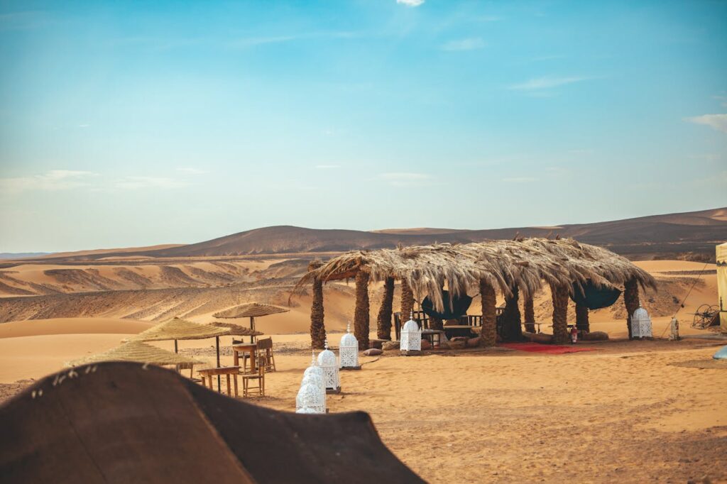 Vue d'un campement dans le désert marocain avec une structure en paille et des palmiers, entourée de tables en bois, de lanternes blanches et de dunes de sable doré sous un ciel bleu clair et des collines brunes au loin