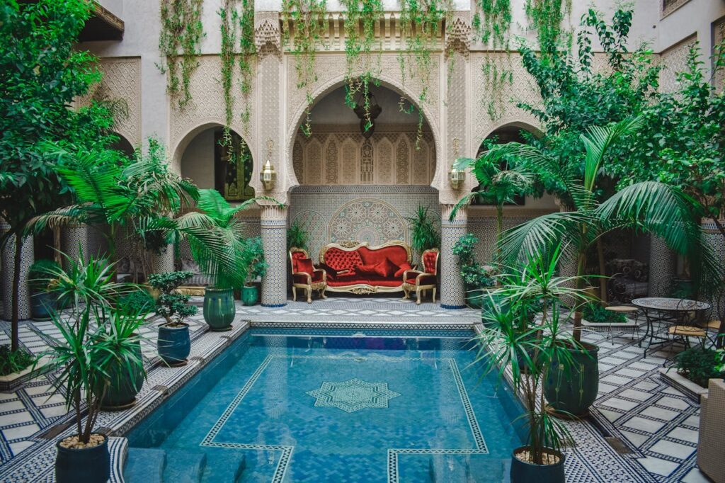 Ce splendide riad, niché au cœur de la médina, incarne toute la magie de l’art de vivre marocain. Son patio central dévoile une piscine aux reflets turquoise, bordée de carreaux zelliges finement agencés. Une végétation luxuriante — palmiers, plantes grimpantes et arbustes — enveloppe l’espace d’une atmosphère apaisante. En toile de fond, de majestueuses arcades sculptées et des colonnes décorées de mosaïques mettent en valeur un salon oriental, meublé de fauteuils en velours rouge et d’un canapé au design raffiné. Une oasis de sérénité, idéale pour se détendre après l’effervescence des ruelles de Marrakech.