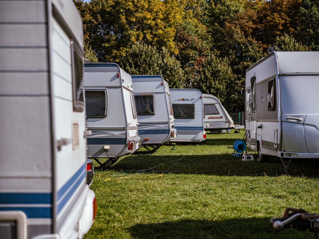 Sur cette image, on voit plusieurs camping-cars (ou caravanes) alignés dans un espace vert, probablement un terrain de camping. L'herbe est bien verte et la lumière indique une belle journée ensoleillée. À l’arrière-plan, une rangée d’arbres aux feuillages mêlés de vert et de teintes automnales (jaunes, orangées) entoure le campement, créant une ambiance paisible.