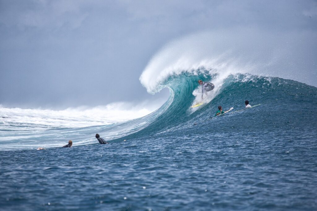 Sur cette image dynamique, on découvre une scène de surf impressionnante en pleine action. Un surfeur est parfaitement positionné dans le tube d’une grande vague turquoise, concentré sur sa trajectoire, tandis que l’eau forme une cavité presque parfaite autour de lui. La vague, puissante et bien formée, déploie une crête blanche éclatante sous un ciel couvert.
À l’avant-plan, plusieurs autres surfeurs attendent leur tour, allongés sur leurs planches ou assis, observant la vague et la performance du surfeur engagé. L’ensemble évoque une ambiance à la fois intense et paisible, typique des meilleurs spots de surf du monde.