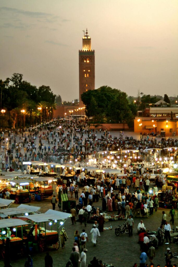 L'image capture une scène animée de la place Jamaa el Fna à Marrakech, au crépuscule. Une foule dense de visiteurs et de locaux se mêle aux stands illuminés