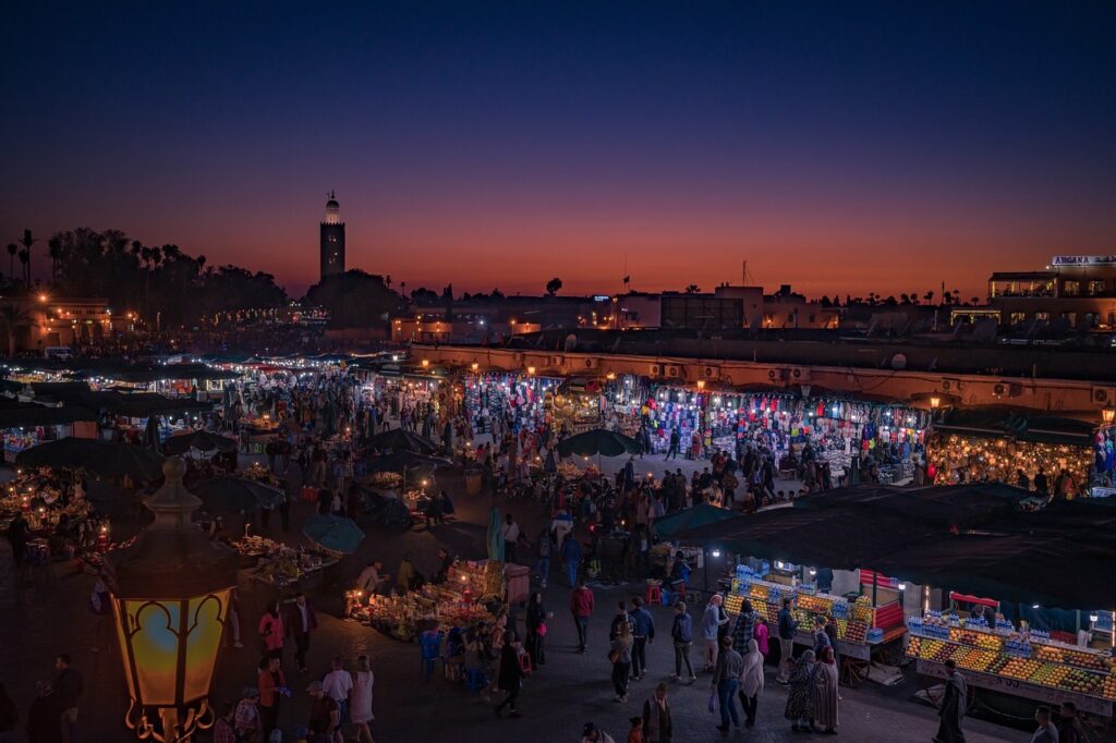L'image capture l'effervescence nocturne de la Médina de Marrakech, baignée dans les teintes chaudes du crépuscule. Une foule dense se presse à travers un dédale de stands illuminés, créant une mosaïque de lumières et d'ombres.