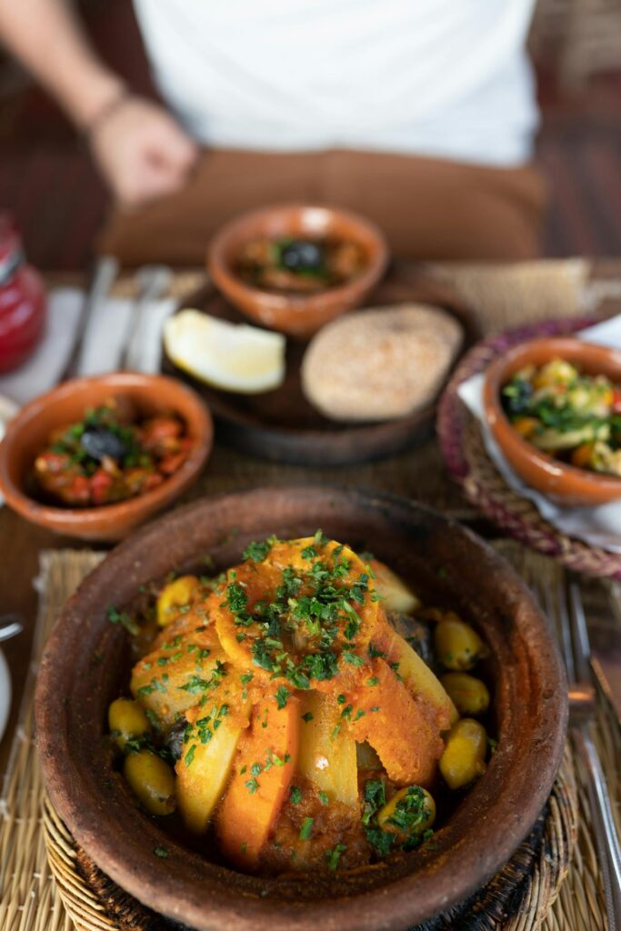 L’image illustre parfaitement la cuisine marocaine, généreuse et colorée. Au premier plan, un tajine de légumes trône dans un plat en terre cuite traditionnel, sublimé par une garniture de carottes, pommes de terre,