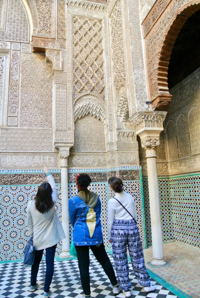 Trois voyageuses émerveillées explorent les somptueuses décorations d’une médersa traditionnelle. Ce type de visite immersive illustre parfaitement l’une des 5 expériences culturelles authentiques à vivre au Maroc,