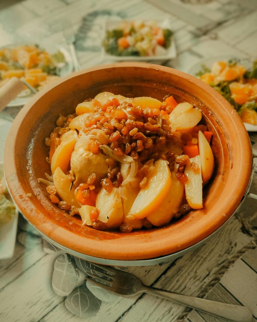 L’image présente un tajine typique de la cuisine marocaine, garni de morceaux fondants de pommes de terre, de carottes, d’oignons et de raisins secs caramélisés, sublimés par des épices douces