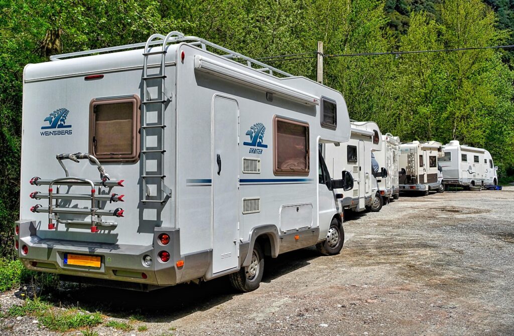 Une rangée de camping-cars, stationne sur un terrain gravillonné entouré de verdure luxuriante. Ces véhicules, prêts pour une aventure Camping-Car Maroc,