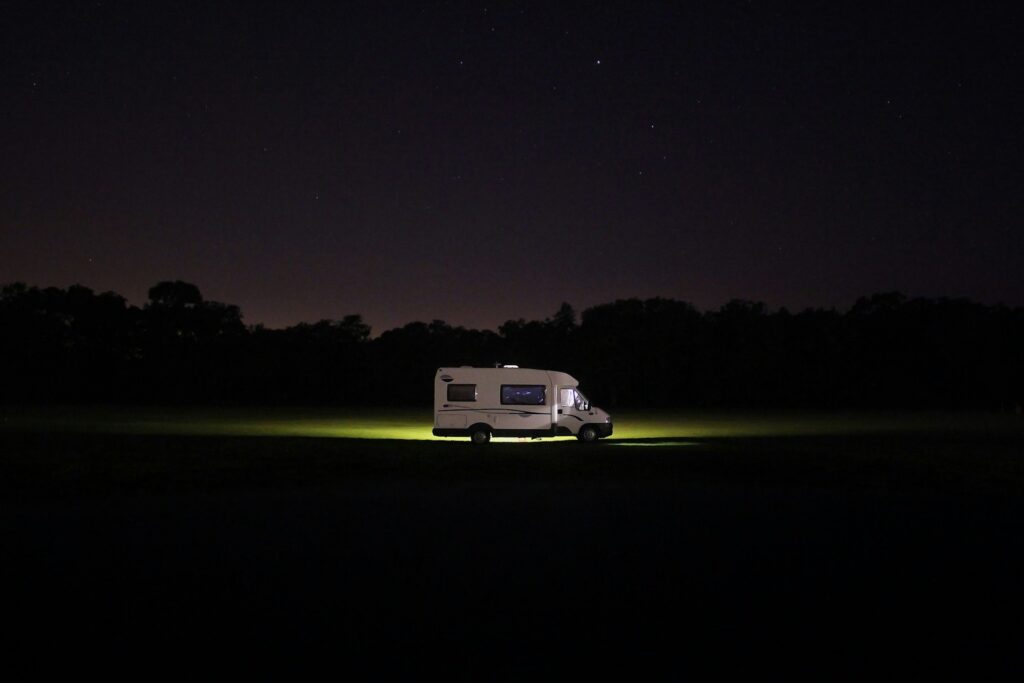 Un camping-car blanc est garé en pleine nature sous un ciel étoilé, éclairé par une lumière douce émanant de ses phares. Ce véhicule incarne une halte paisible idéale pour une aventure Camping-Car Maroc.