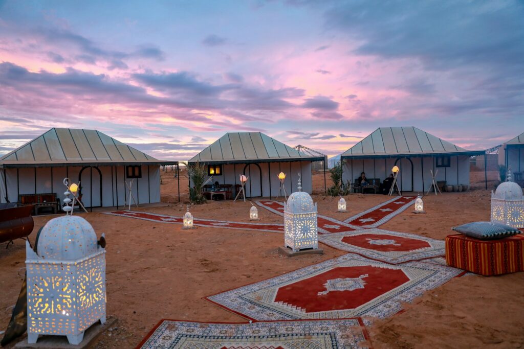 Un campement de Glamping de Luxe au Maroc au coucher du soleil, avec des tentes élégantes bordées de tapis rouges ornés de motifs traditionnels