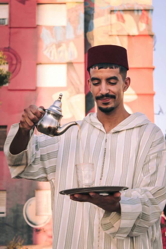 Un homme vêtu d'une djellaba blanche à rayures grises verse du thé à la menthe d'une théière en argent. Ce geste représente l’hospitalité Marocaine