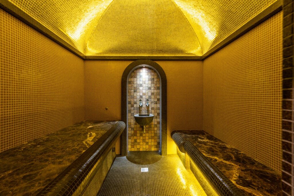 Intérieur d’un hammam marocain traditionnel, aux murs et plafond recouverts de mosaïques dorées et brunes, avec deux banquettes en marbre et un petit bassin encastré sous une arche éclairée