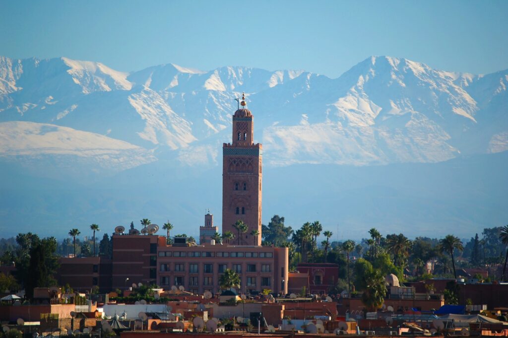 Marrakech photogénique avec la silhouette emblématique de la Koutoubia se détachant sur le panorama majestueux des montagnes de l’Atlas enneigées sous un ciel clair.