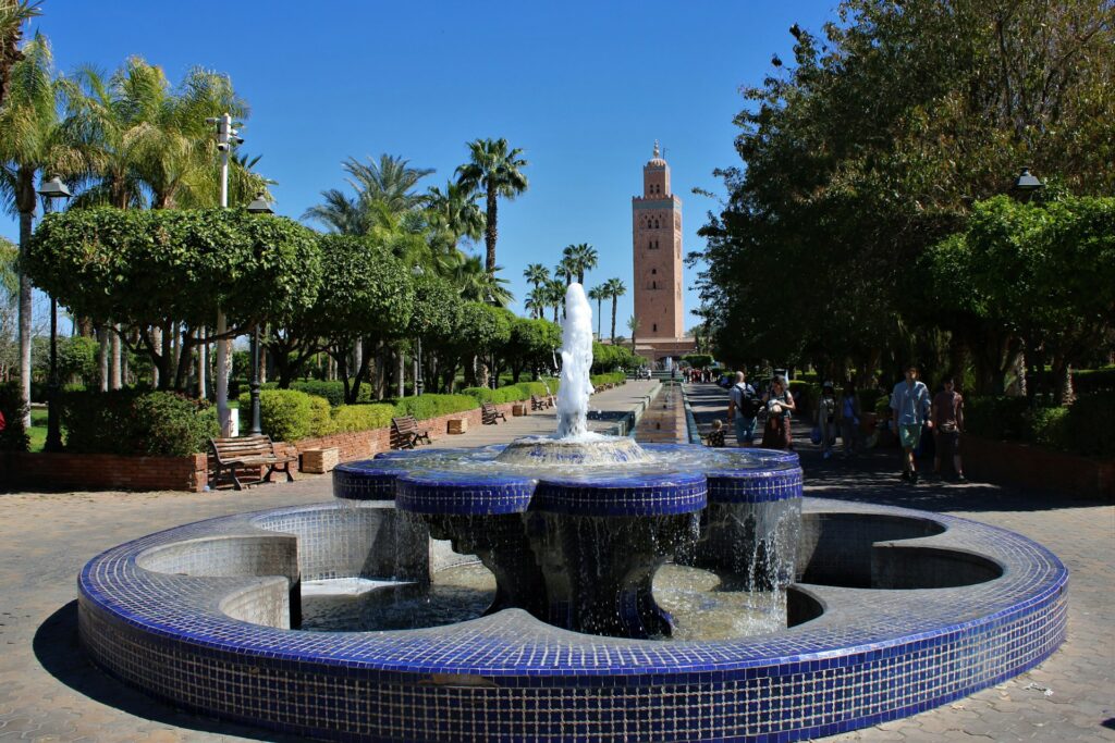 Allée verdoyante du parc de la Koutoubia avec une fontaine en zellige bleu au premier plan et le minaret en arrière-plan, une scène emblématique de Marrakech photogenique sous un ciel parfaitement bleu.