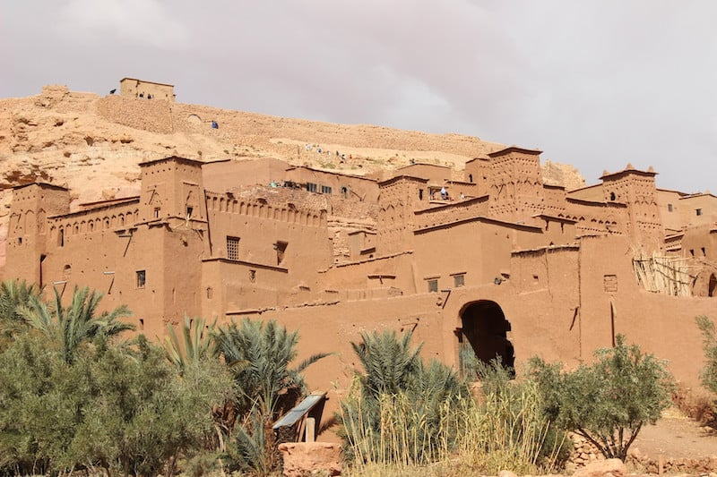 Un ksar (village fortifié) en pisé de couleur ocre rouge. Les bâtiments cubiques avec de petites fenêtres et des créneaux sont étagés sur une colline aride. Il reflète un Maroc authentique : expériences culturelles et immersion locale.